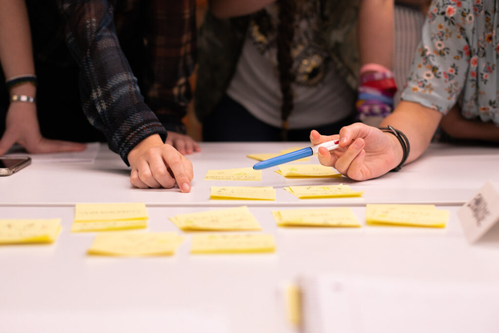 A close-up of post-it notes on a table. A group of students use the post-it notes to explore solutions to a real business problem.